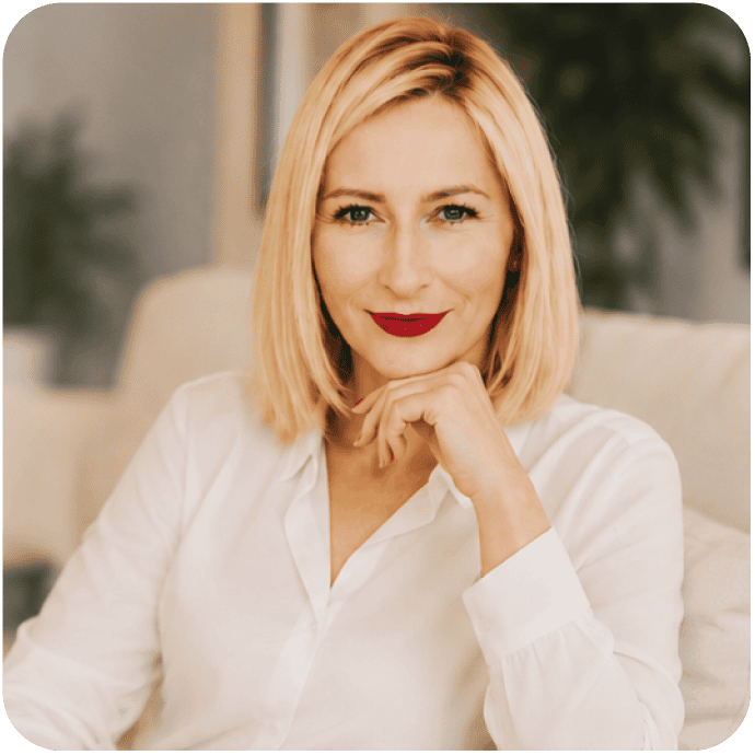 Krystyna Wójcik -Kobieta o prostych blond włosach i z czerwoną szminką siedzi na kanapie w białej bluzce, opierając brodę na dłoni i patrząc w obiektyw z neutralnym wyrazem twarzy.