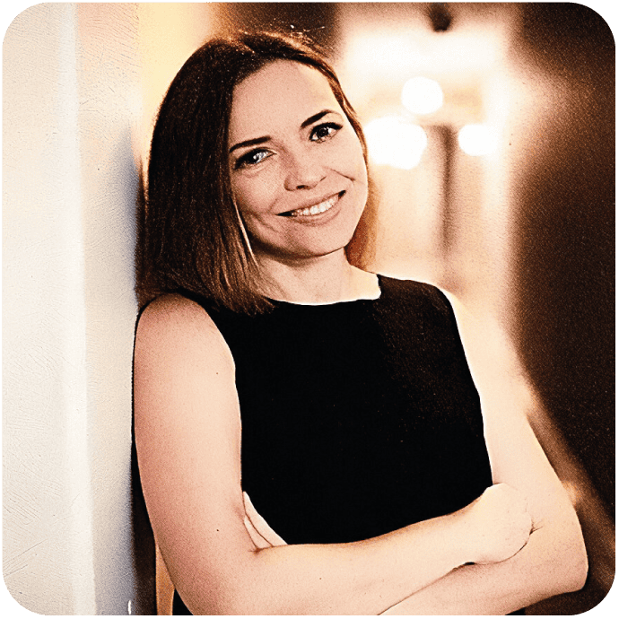 Monika Trypuz -Kobieta o prostych brązowych włosach, ubrana w czarny top bez rękawów, stoi z założonymi rękami, uśmiechając się i opierając się o jasną ścianę w delikatnie oświetlonym korytarzu.