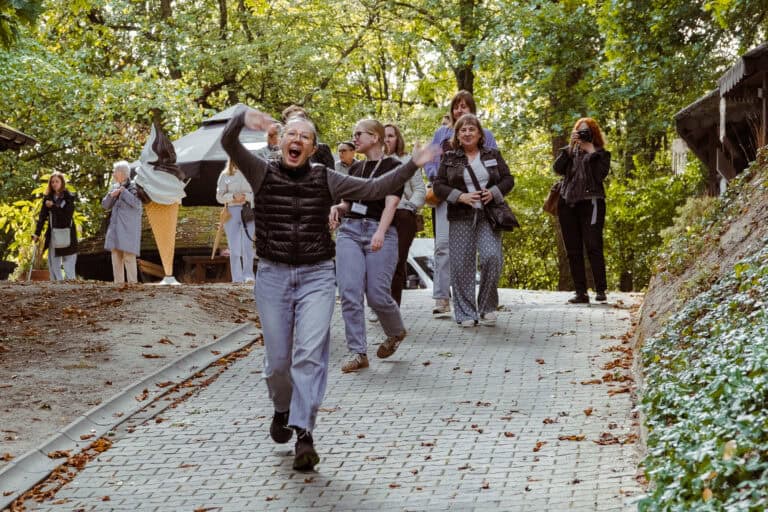 Grupa ludzi idzie na świeżym powietrzu po utwardzonej ścieżce otoczonej drzewami. Osoba idąca z przodu biegnie i unosi ręce, podczas gdy pozostali idą i uśmiechają się w tle.