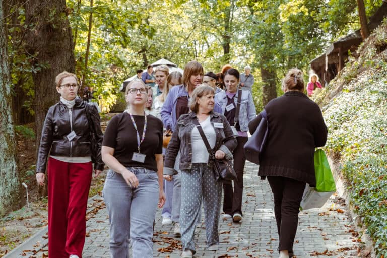 W słoneczny dzień grupa ludzi idzie wzdłuż wysadzanej drzewami ścieżki; niektórzy mają przypięte plakietki z imionami i niosą torby.