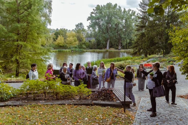 Grupa ludzi stoi na ścieżce w parku w pobliżu stawu, w otoczeniu drzew i zieleni; niektórzy trzymają aparaty fotograficzne i robią zdjęcia.