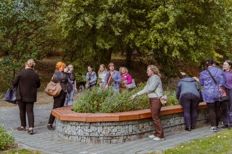 Grupa ludzi stoi i gromadzi się wokół okrągłej, podwyższonej grządki w parku, otoczonej zielonymi drzewami.