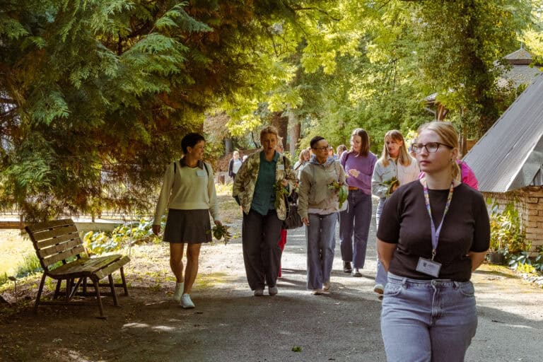 Grupa kobiet idzie zacienioną ścieżką w parku; niektóre trzymają liście, a przed nimi idzie inna kobieta z zawieszką na szyi.
