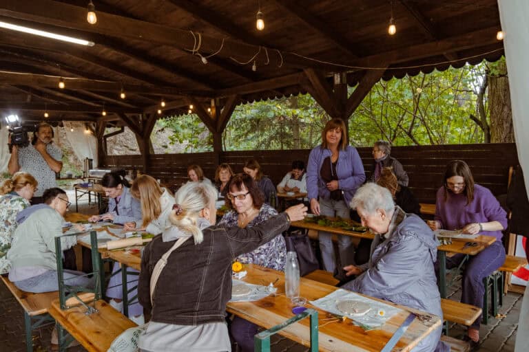 Grupa ludzi siedzi przy drewnianych stołach pod altaną, zajmując się rękodziełem z wykorzystaniem roślin i kwiatów; w tle ktoś kręci film.