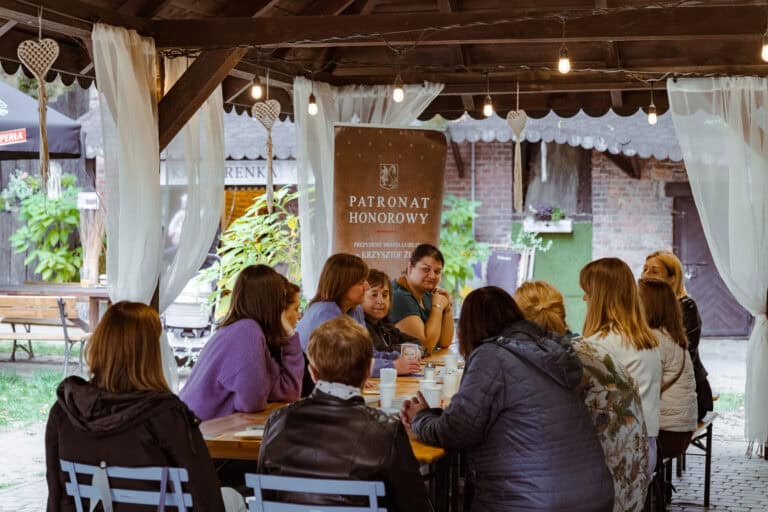 Grupa ludzi siedzi przy stole pod altaną i prowadzi rozmowę. Scena wydaje się rozgrywać na świeżym powietrzu, a w tle widać banery i zieleń.