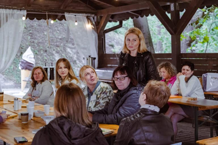 Grupa kobiet siedzi przy drewnianych stołach pod altaną i słucha kobiety, która stoi i przemawia. Na stołach leżą kubki, kartki i torby. Na zewnątrz widać drzewa oraz dużą dekorację w kształcie rożka do lodów.