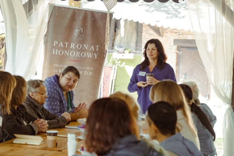 Kobieta stoi i przemawia do siedzącej grupy przy stoliku na świeżym powietrzu, obok transparentu z napisem „Patronat Honorowy”. Kilka osób jej słucha, a niektóre mają przed sobą filiżanki z kawą.