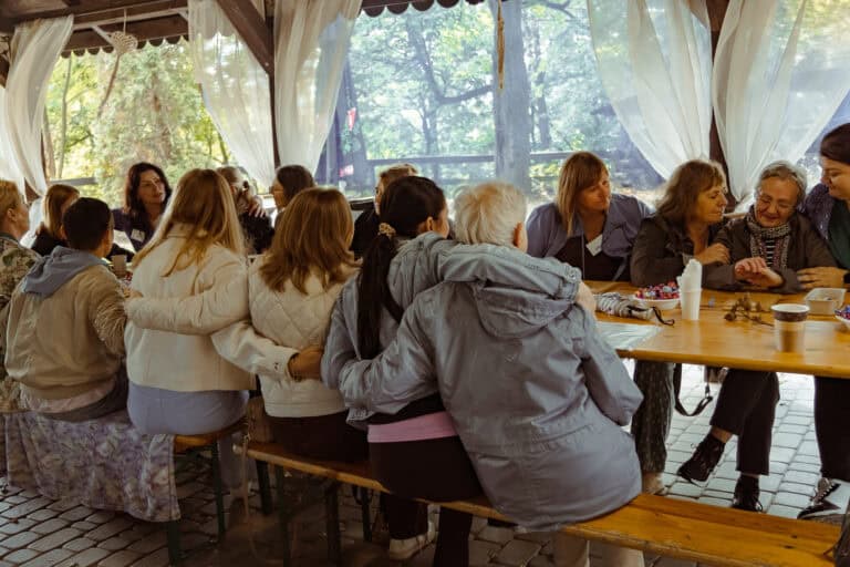 Grupa ludzi siedzi blisko siebie przy długim drewnianym stole piknikowym pod markizą, rozmawiając i popijając napoje na świeżym powietrzu.