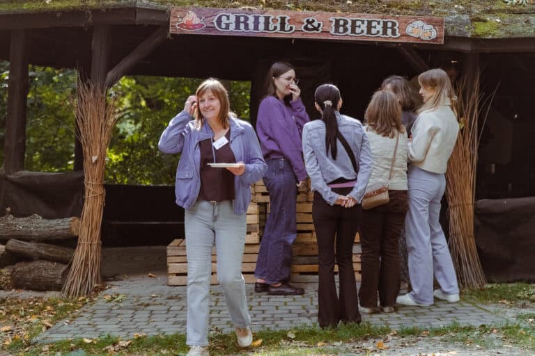 Grupa kobiet stoi i rozmawia przed rustykalnym napisem „Grill & Beer”; jedna z nich odchodzi z uśmiechem, trzymając talerz.