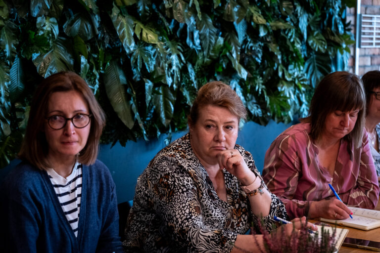 Trzy kobiety siedzące przy stole przed ścianą z zielonymi roślinami; jedna z nich pisze w notatniku.