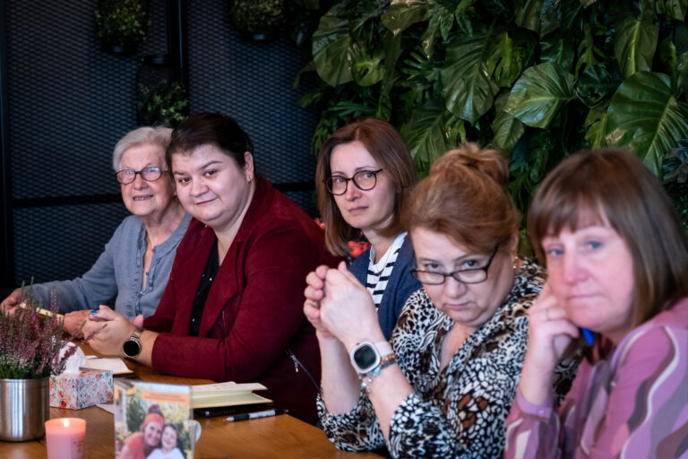 Pięć kobiet siedzących razem przy stole przed ścianą porośniętą roślinnością.