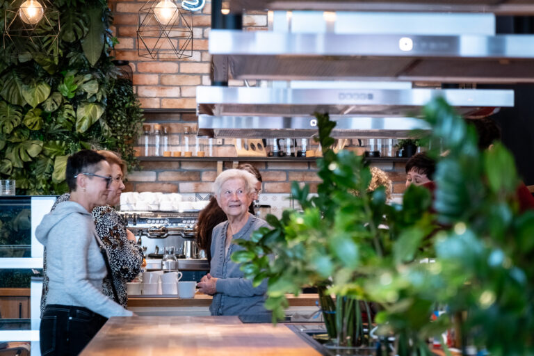 Trzy kobiety stoją przy ladzie kawiarni, a w tle widać ekspres do espresso.