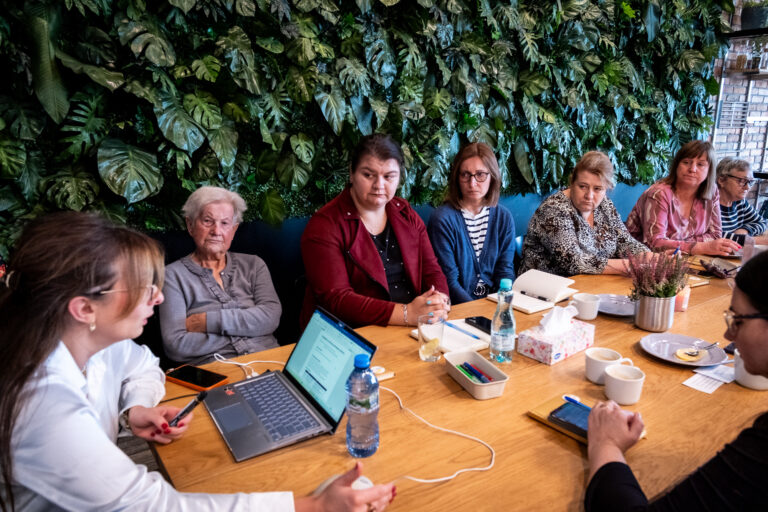 Grupa kobiet siedzi przy długim drewnianym stole podczas spotkania przed ścianą pokrytą zielonymi roślinami.