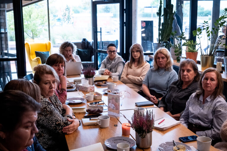 Grupa osób siedzi przy długim drewnianym stole podczas spotkania w nasłonecznionym pomieszczeniu.
