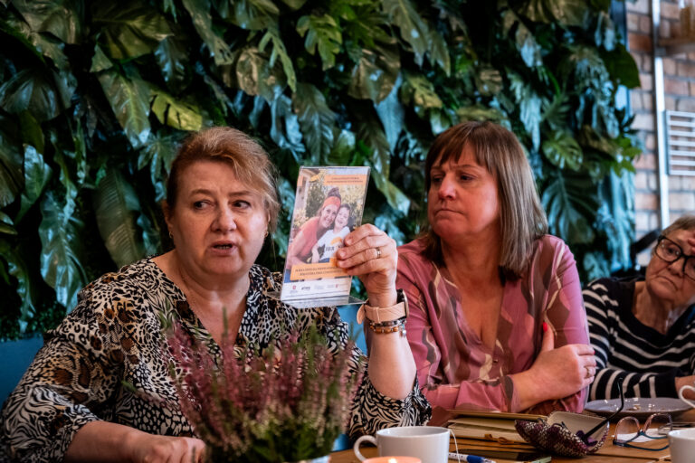 Kobieta trzyma w ręku małą ulotkę informacyjną ze zdjęciem kobiety i dziecka, siedząc przy stole wraz z dwiema innymi kobietami.