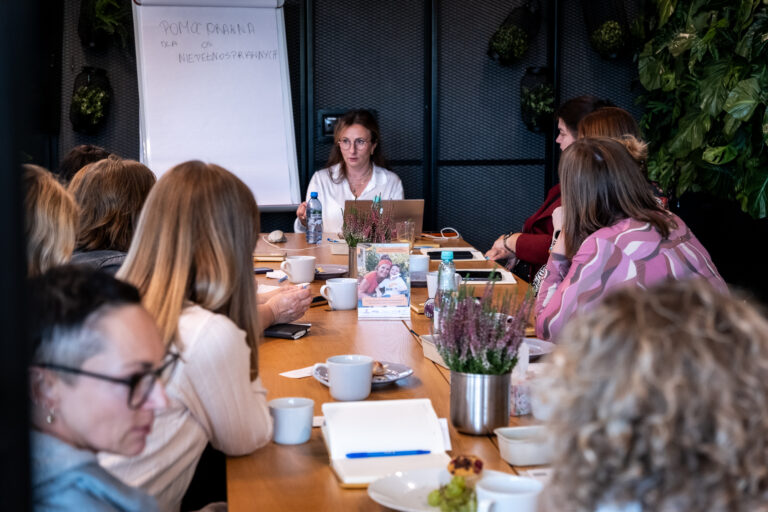 Kobieta prowadzi spotkanie grupowe przy długim stole, a w tle widać tablicę, na której widnieje napis „Pomoc prawna dla osób niepełnosprawnych”.
