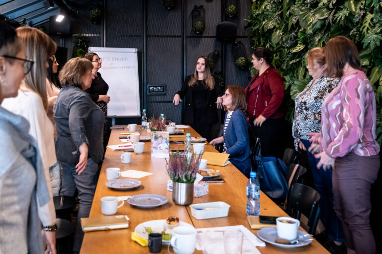 Podczas spotkania w sali z „żywą ścianą” grupa kobiet stoi wokół długiego stołu konferencyjnego.