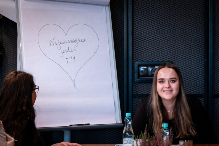 Kobieta uśmiecha się przy stoliku obok tablicy, na której widnieje narysowane ręcznie serce wraz z napisem w języku polskim: „Najważniejsza jesteś TY”.