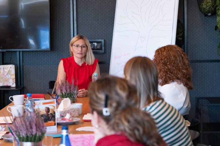 Kobieta w czerwonej bluzce prowadzi spotkanie z grupą osób siedzących przy stole, na którym znajduje się tablica z ręcznie narysowanym schematem drzewa.