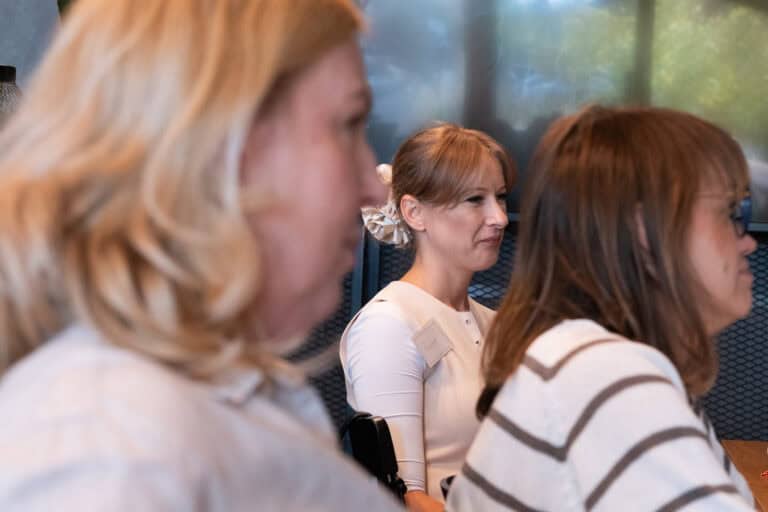Trzy osoby siedzące przy stole podczas spotkania; osoba pośrodku ma przypiętą plakietkę z imieniem.