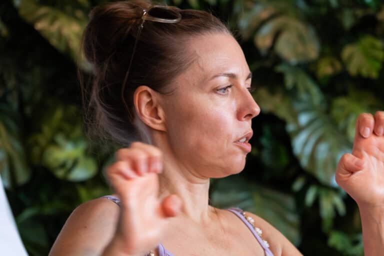 Kobieta z włosami upiętymi w kok gestykuluje obiema rękami przed ścianą pokrytą bujną zielenią.