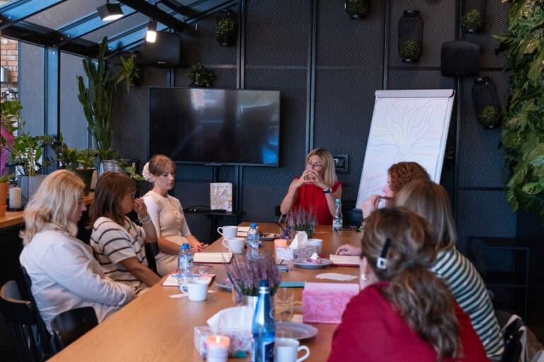 Kobieta prowadzi spotkanie grupy kobiet siedzących przy stole konferencyjnym, obok którego znajduje się tablica suchościeralna.